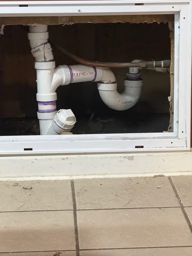 An opened access panel under a kitchen sink with a new PVC trap and cleanout plug, finished by Peterson