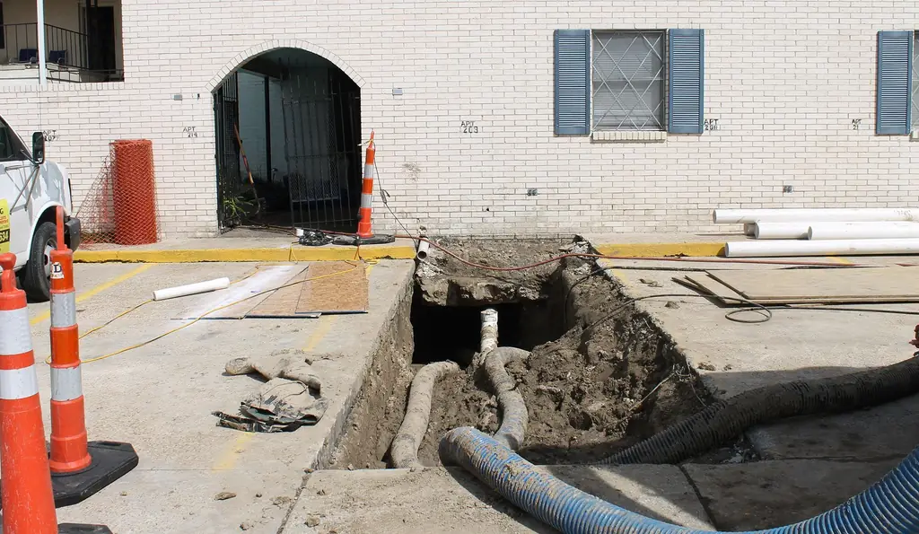 Open trench cut through a parking-lot slab beside a brick apartment building, vac hoses running into it